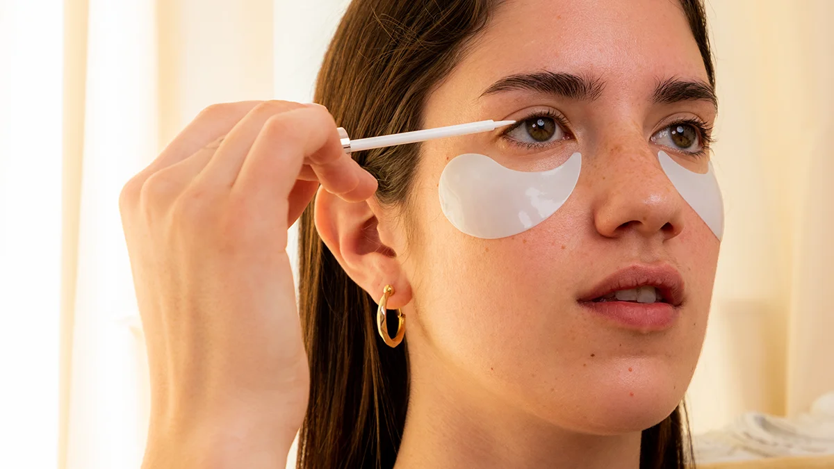 A young woman with undereye patches applying IRIS eyelash serum without prostaglandin